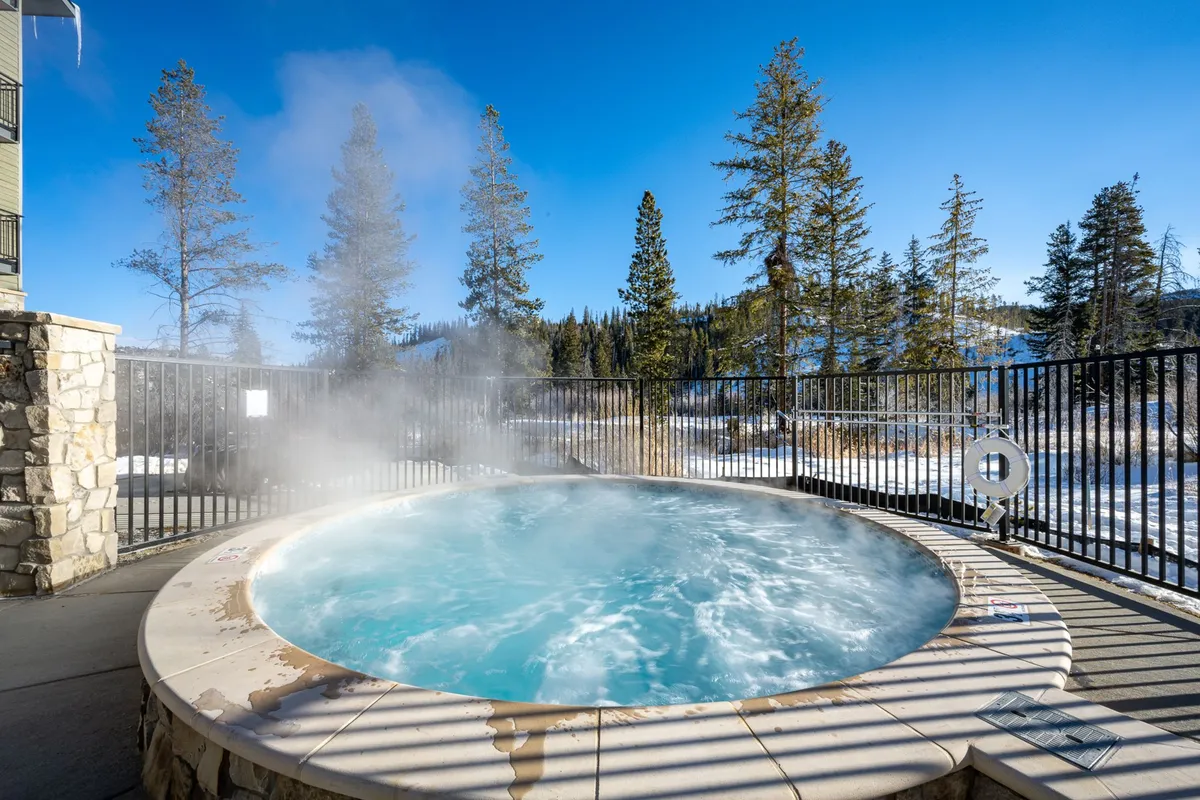 An inviting outdoor community hot tub surrounded by natural landscaping, with lush trees and a dramatic mountain backdrop. The scene captures guests relaxing and socializing in the steaming water, with the golden light of sunset adding a tranquil, rejuvenating mood.