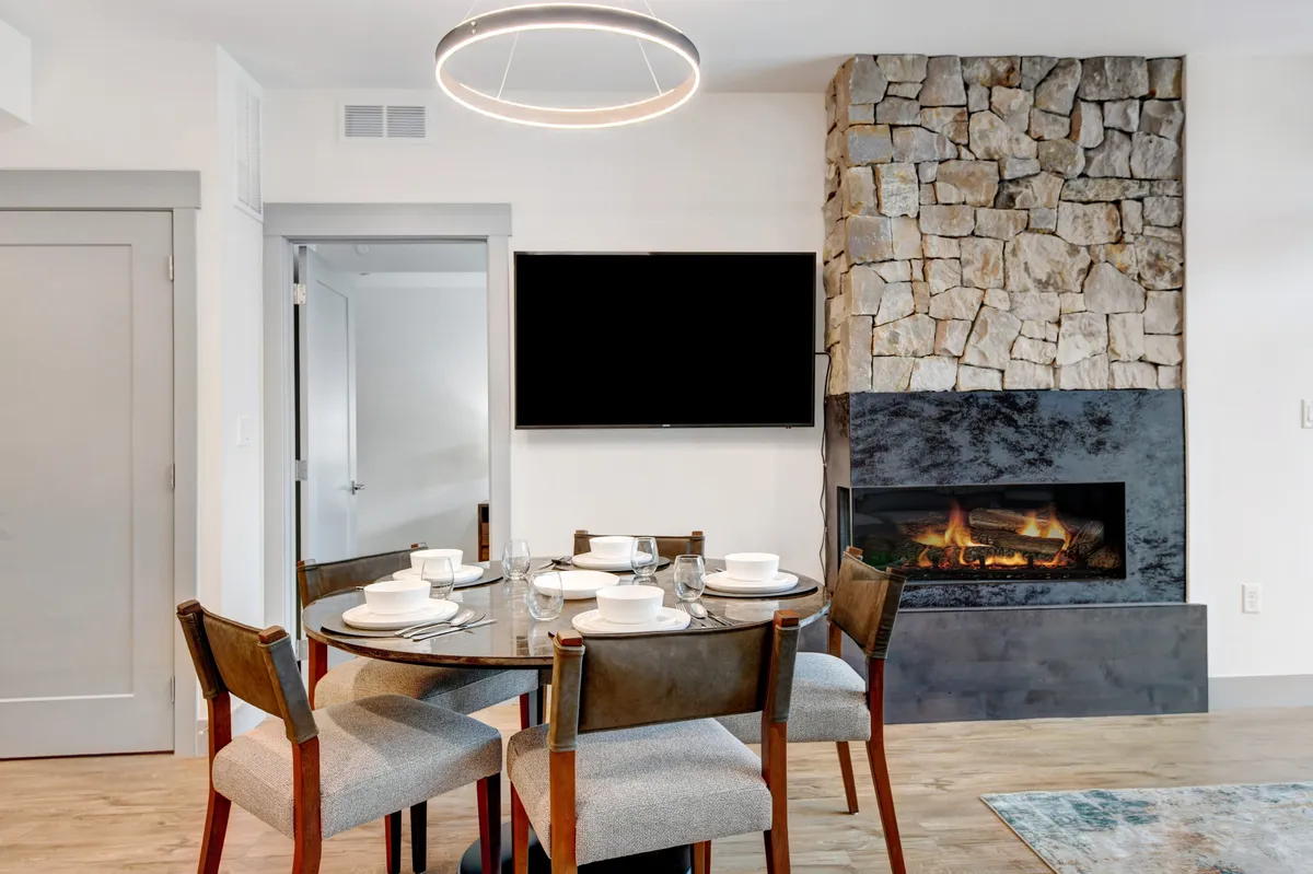 A warm and modern living/dining area with a stone fireplace, wall-mounted TV, and an elegant dining table set for gathering. The space is ideal for family meals, intimate gatherings, and enjoying entertainment together.