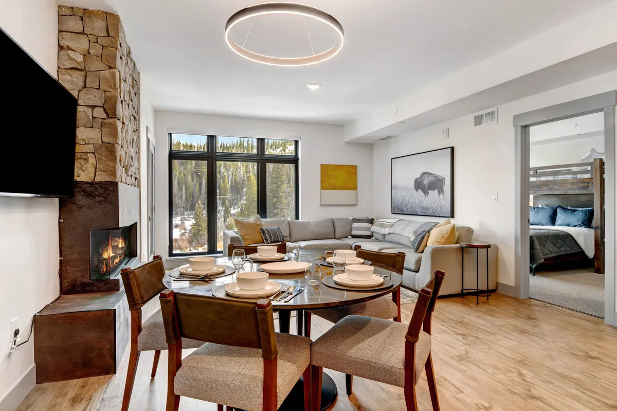A dining area with a round table set for a meal is featured, accompanied by a stone fireplace, large windows, and a comfortable sectional sofa. The ambiance is warm, suggesting shared meals, laughter, and a connection to the living area, as described in the section.