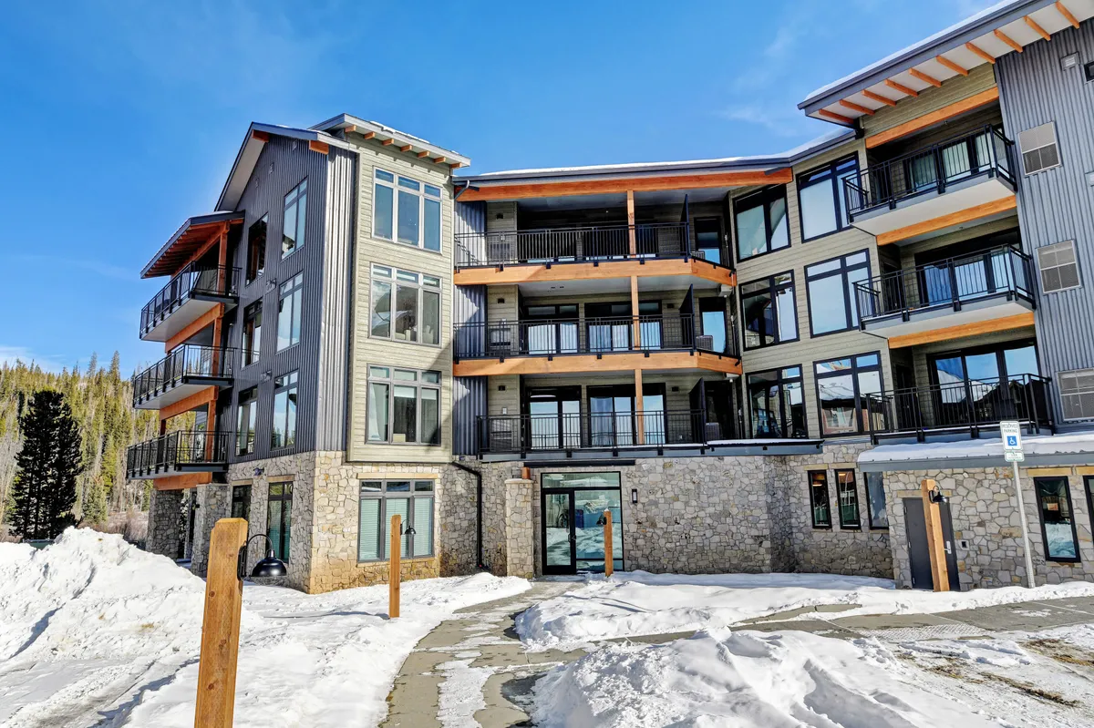 A welcoming exterior view of the condo building showcasing modern architecture with large windows and a stone facade. Snow blankets the ground, and balconies overlook the surrounding landscape, suggesting both comfort and a connection to the outdoors.