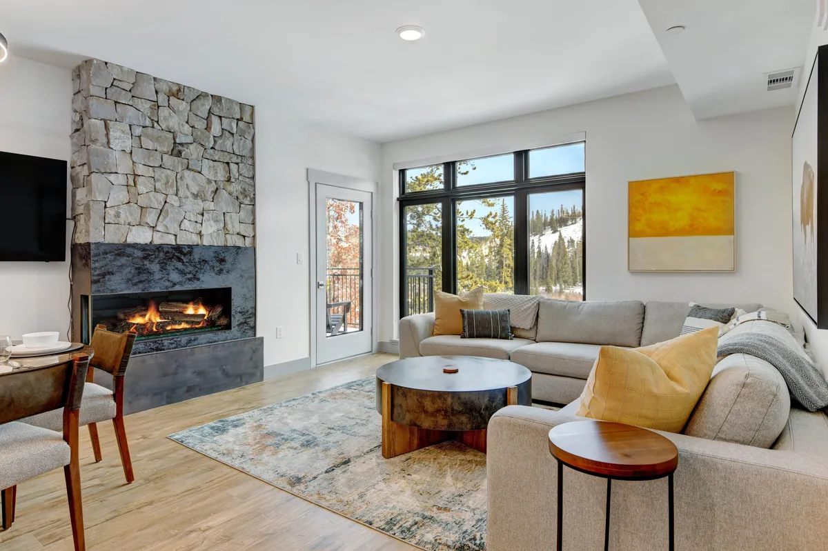 The image showcases a stylish and inviting living space featuring a grand stone fireplace, plush sofa, striking artwork, and expansive windows that frame stunning mountain views. Natural light fills the room, highlighting a cozy dining area perfect for shared meals or game nights, while the snowy landscape outside adds to the warmth and ambiance inside.