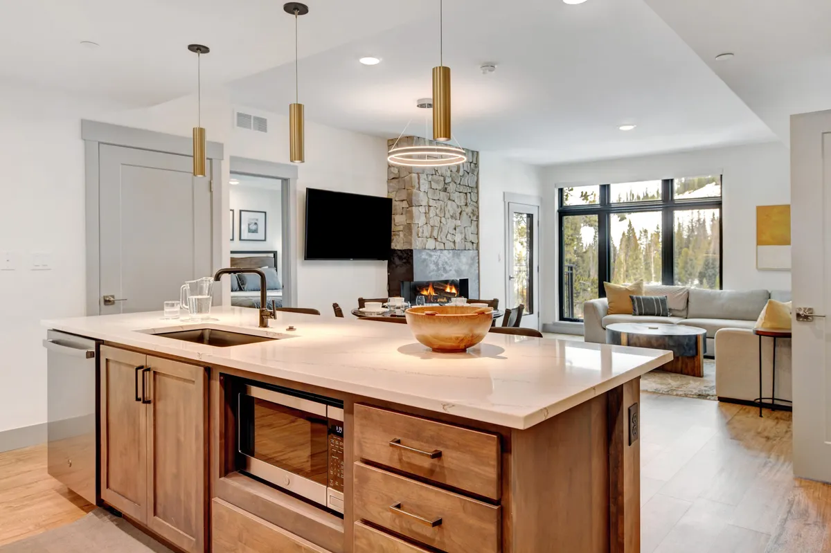 A spacious open-concept living area featuring a cozy seating arrangement in front of a modern fireplace, with a kitchen island and large windows that frame views of the surrounding forest. The setup emphasizes togetherness, comfort, and seamless flow between living, dining, and kitchen spaces.