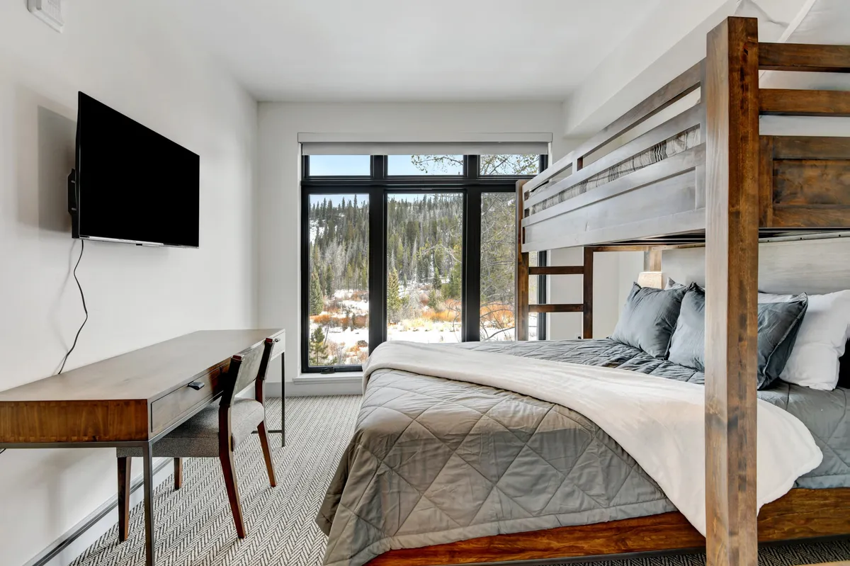 A cheerful bunk room with a twin-over-queen bunk bed, a functional desk and chair setup for work or study, and a large window providing scenic mountain views. The space exudes warmth and is tailored for both young children and families, offering a TV and ample room for play or productivity.
