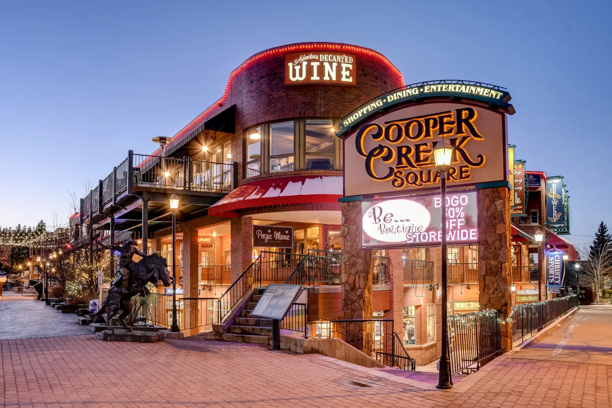 A lively exterior shot featuring the Cooper Creek Square sign, festive decorative lighting, and visible signage for a wine shop. The scene showcases a vibrant shopping and dining area bustling with energy, ideal for social gatherings, events, and casual outings.